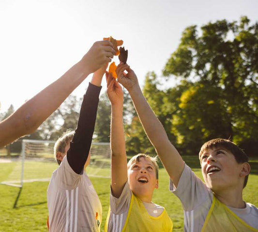 Three young soccer players in jerseys reach up together to share a piece of waffle on a sunlit field with a goal and trees behind them
