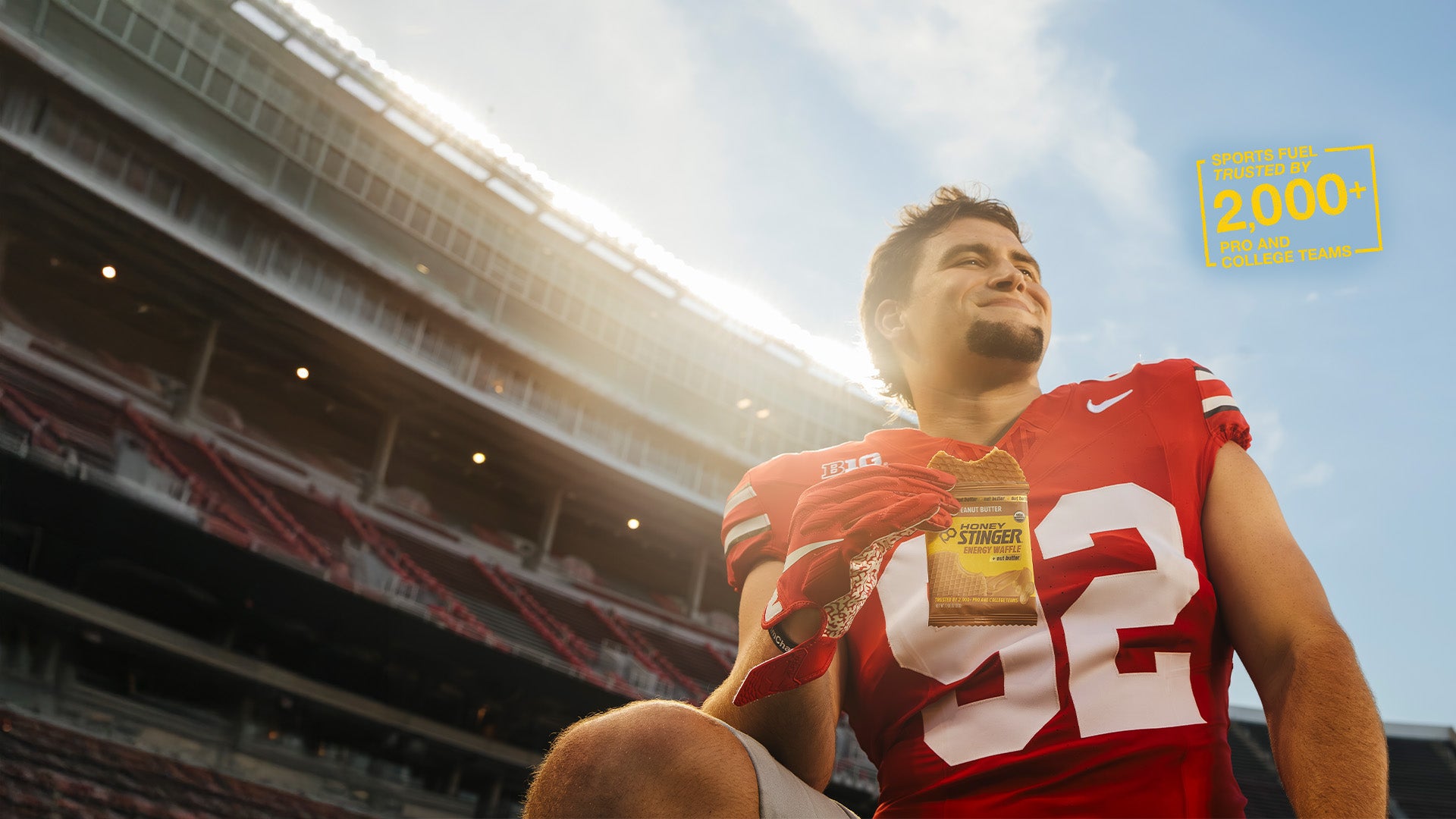 Person in a red football jersey holding a Honey Stinger Peanut Butter Energy Waffle against a stadium background
