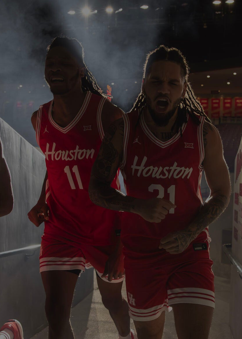 Two basketball players in red Houston jerseys run through a tunnel, with team names and years printed in red on the wall to their right.
