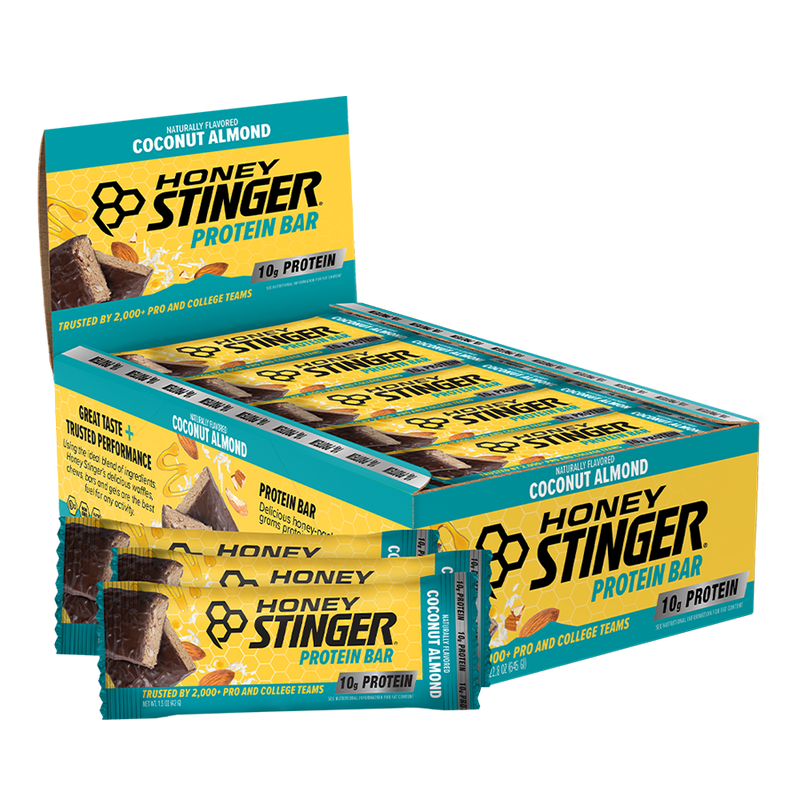 A box of Honey Stinger Protein Bars in Coconut Almond flavor, with several individual bars wrapped in teal and yellow packaging displayed in front on a white background.