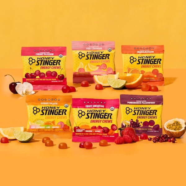 Six Honey Stinger Energy Chews packets (Sampler Pack flavors) standing upright on a yellow/orange background surrounded by cherries, lemon, and other fruit